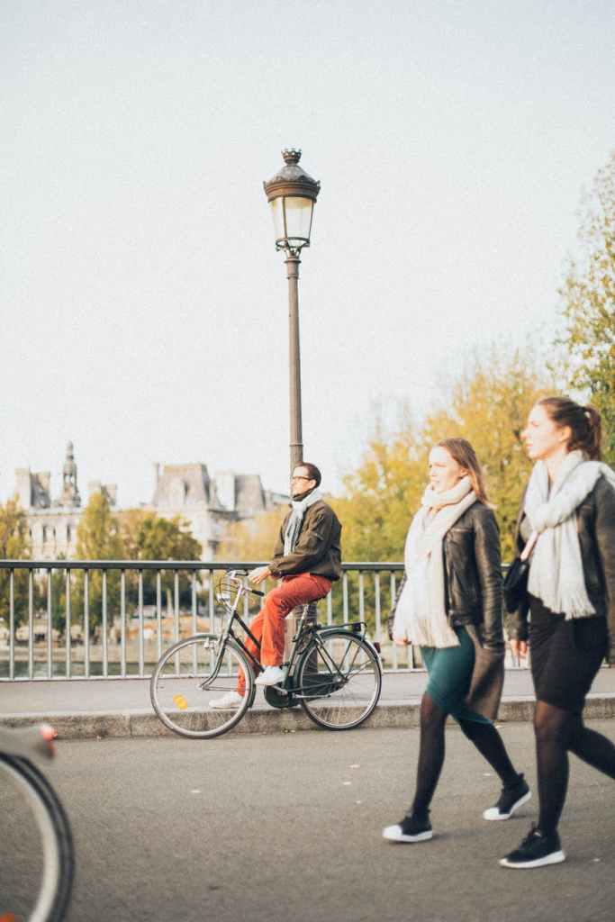 A man dressed casually sits on his bike, foot on the pavement, on his ride around town. Two young women pedestrians pass him by.