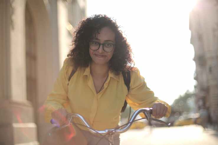 A smiling woman pushing her bicycle on a sunny day amongst some tall buildings